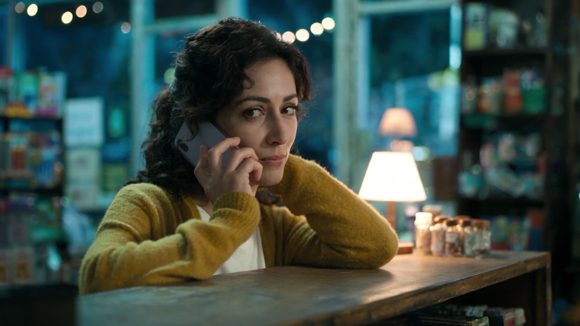 A woman in a mustard cardigan holds a phone to her ear at a shop counter at night.